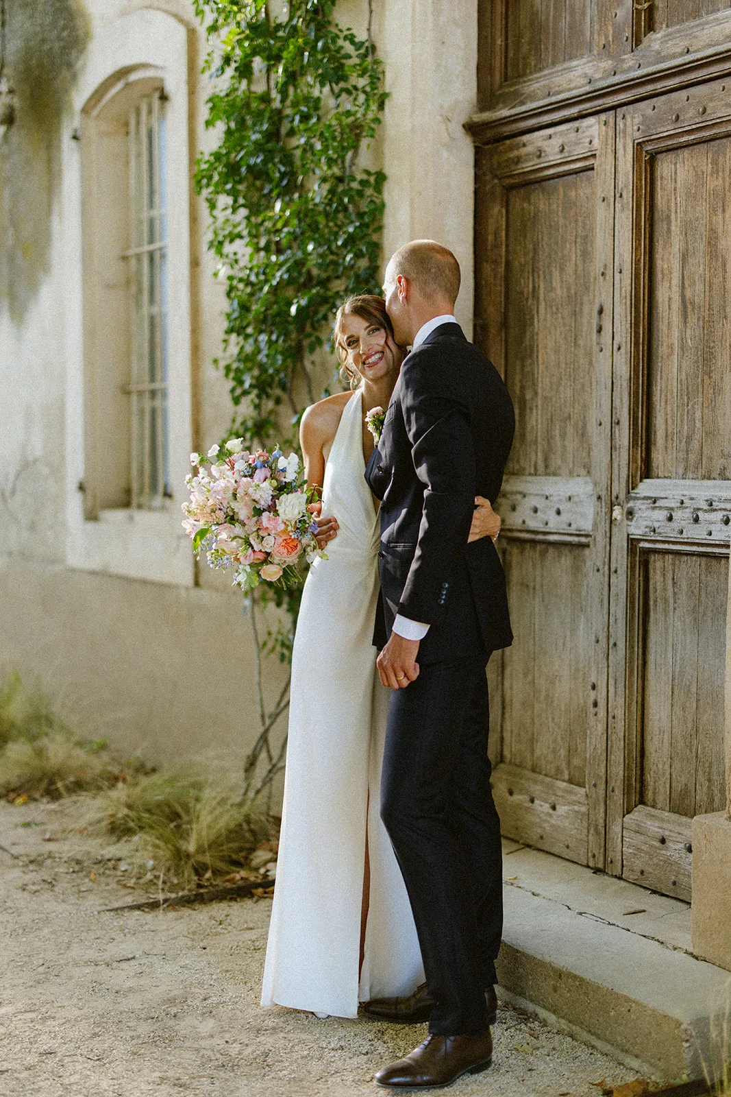 Réka et Malo enlacés devant la grande porte en bois du Château du Griffon, bouquet de fleurs pastel