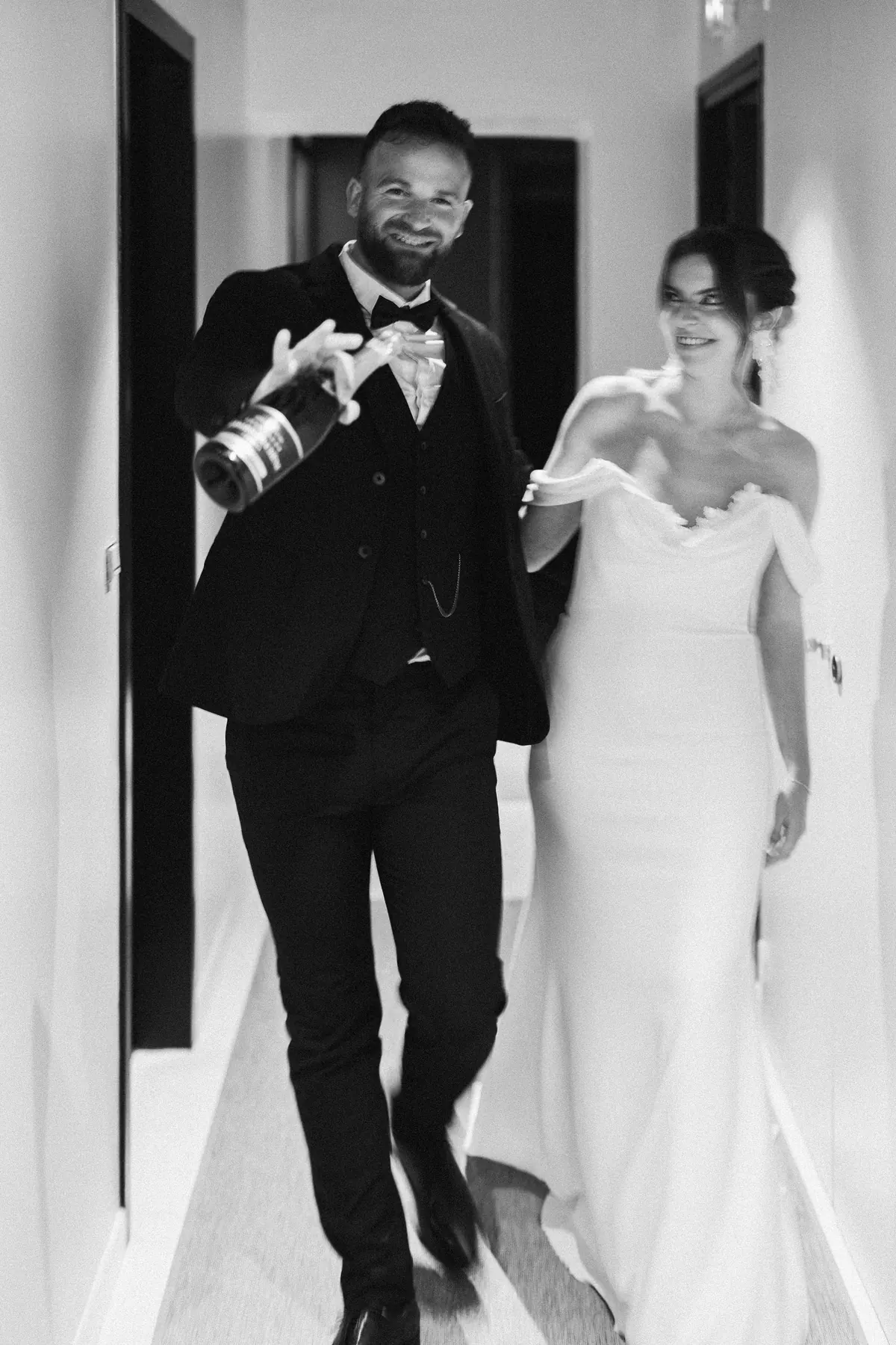 Mariés riant dans un couloir, le marié tenant une bouteille de champagne, photo en noir et blanc