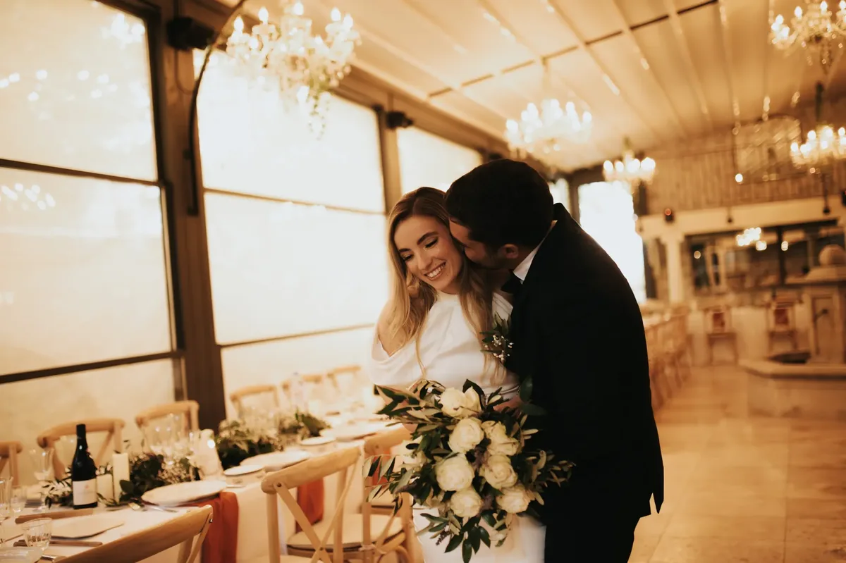 Mariée souriante enlacée par son époux qui l'embrasse sur la joue, bouquet de roses blanches au premier plan, salle de réception aux lustres en cristal